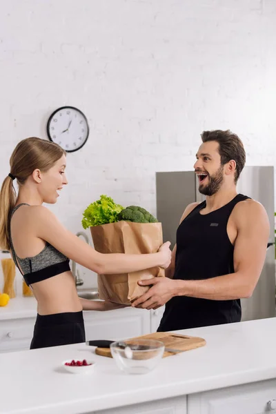 Chica sorprendida sosteniendo bolsa de papel con comestibles cerca del hombre barbudo en la cocina - foto de stock