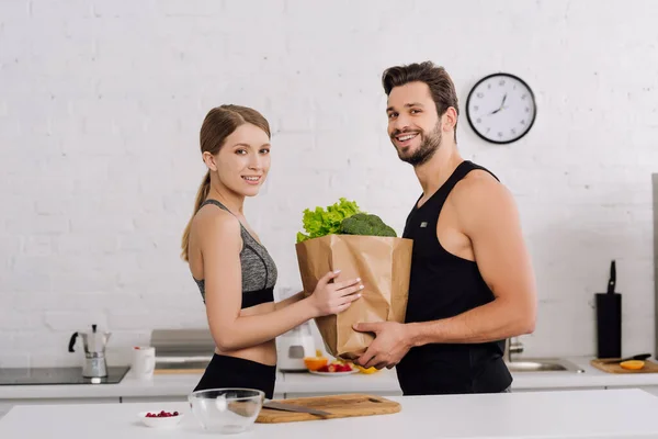 Chica feliz y barbudo hombre sosteniendo bolsa de papel con comestibles en la cocina - foto de stock