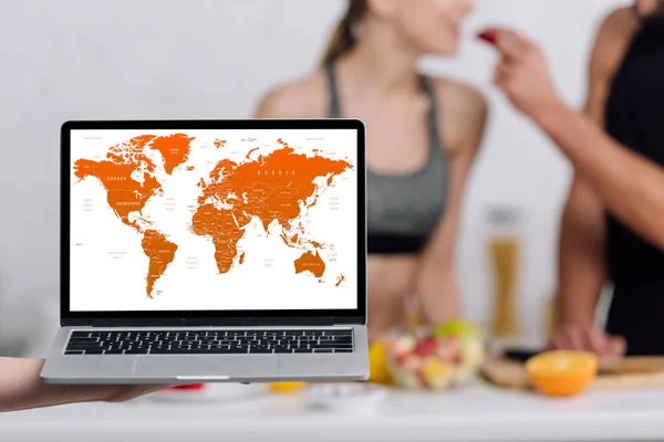 Foco selectivo de la computadora portátil con mapa del mundo en la pantalla cerca de la pareja en la cocina - foto de stock