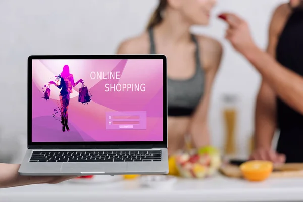 Enfoque selectivo de la computadora portátil con el sitio web de compras en línea en la pantalla cerca de pareja en la cocina - foto de stock