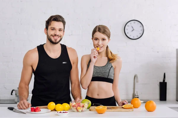 Felice uomo sorridente vicino donna mangiare affettato arancione in cucina — Foto stock