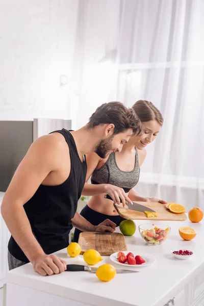 Hombre feliz mirando a la mujer cocinar ensalada de frutas - foto de stock