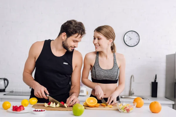 Alegre pareja sonriendo mientras se prepara ensalada de frutas - foto de stock