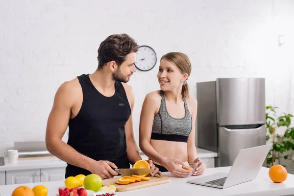 Happy girl looking at sportive man near laptop and fruits in kitchen — Stock Photo