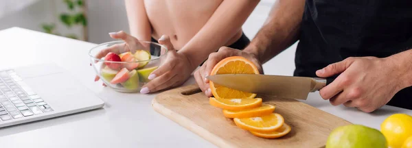 Plano panorámico del hombre cortando naranja cerca de la mujer, ensalada y portátil - foto de stock