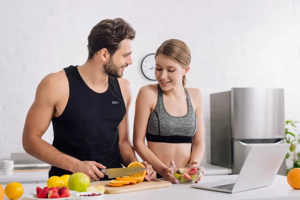 Счастливая девушка с салатом рядом спортивный человек, ноутбук и фрукты на кухне — стоковое фото