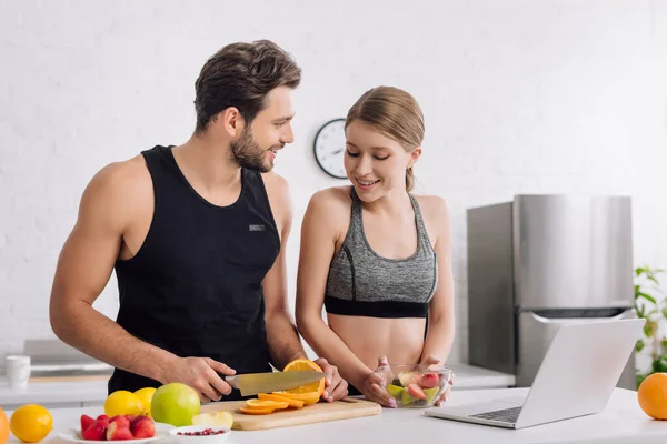 Fille heureuse avec salade près de l'homme sportif, ordinateur portable et fruits dans la cuisine — Photo de stock