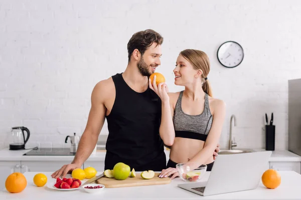 Happy girl with orange near sportive man, laptop and fruits in kitchen — Stock Photo