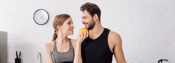 Panoramic shot of happy girl holding orange and looking at cheerful man — Stock Photo