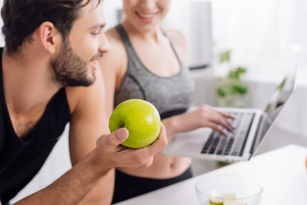 Selektiver Fokus eines glücklichen Mannes, der einen Apfel in der Hand hält und auf einen Laptop neben einer Frau blickt — Stockfoto