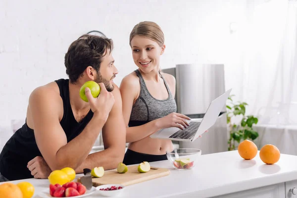 Счастливый мужчина с яблоком смотрит на женщину с ноутбуком рядом с фруктами — стоковое фото