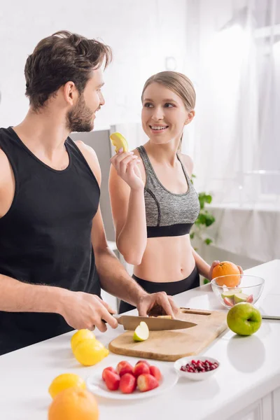 Hombre feliz cortando manzana cerca de la mujer en ropa deportiva - foto de stock