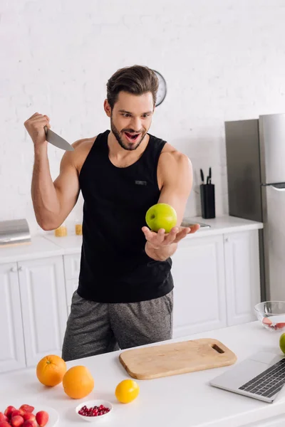 Hombre guapo con cuchillo sosteniendo manzana cerca de frutas y portátil - foto de stock