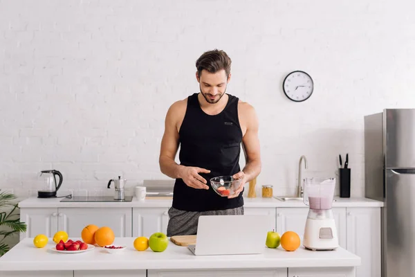 Schöner Mann schaut auf Laptop neben leckeren Früchten und Mixer mit Smoothie — Stockfoto