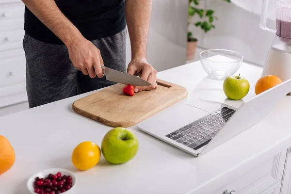 Abgeschnittene Ansicht eines Mannes, der Erdbeeren in der Nähe von Laptop schneidet — Stockfoto