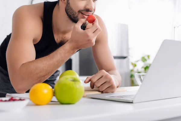 Cropped view of happy man using laptop and eating strawberry — Stock Photo