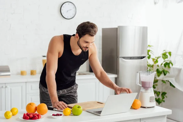 Bell'uomo che utilizza il computer portatile vicino frullatore con frullato e frutta — Foto stock