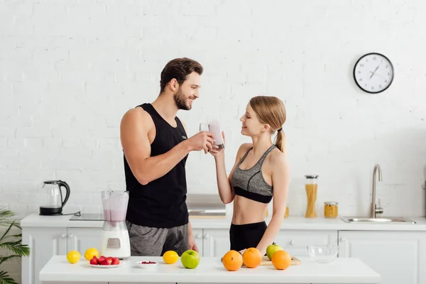 Side view of cheerful man and woman clinking glasses with smoothie near fruits — Stock Photo