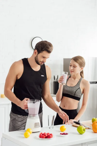 Homem bonito segurando liquidificador perto menina esportiva com vidro de smoothie — Fotografia de Stock