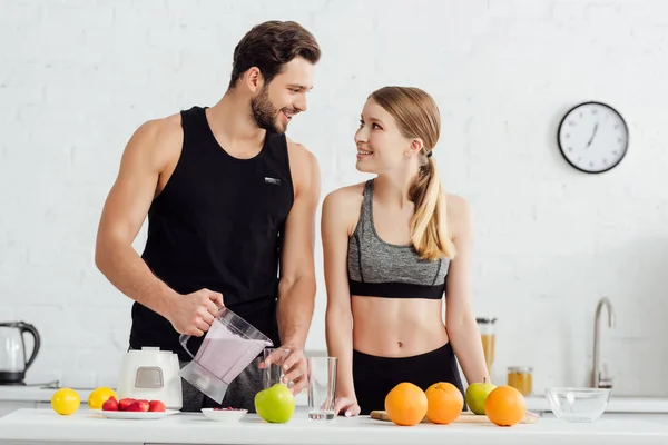 Homem esportivo derramando smoothie em vidro perto de mulher feliz e frutas — Fotografia de Stock