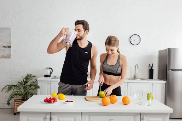 Feliz chica corte manzana cerca hombre bebiendo smoothie de licuadora - foto de stock