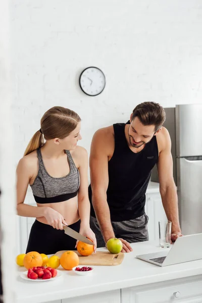 Sportive girl cutting orange near man and laptop — Stock Photo