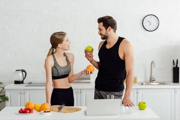 Feliz hombre y mujer con sabrosas frutas mirándose el uno al otro cerca del portátil - foto de stock