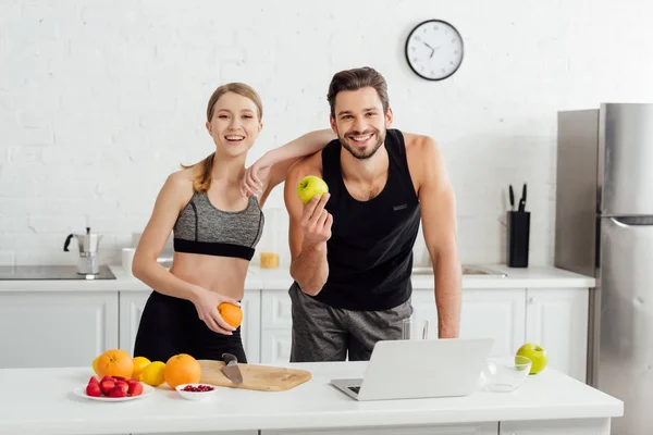 Glücklicher Mann und Frau mit leckeren Früchten, die in Laptopnähe in die Kamera schauen — Stockfoto