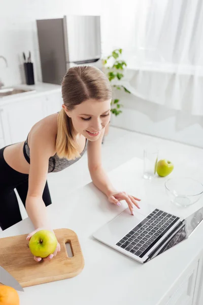 Счастливая и спортивная женщина держит вкусное яблоко и с помощью ноутбука — стоковое фото
