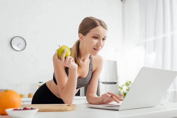 Enfoque selectivo de la mujer deportiva sosteniendo sabrosa manzana y el uso de ordenador portátil - foto de stock