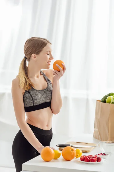 Mulher feliz e esportiva cheirando laranja na cozinha — Fotografia de Stock