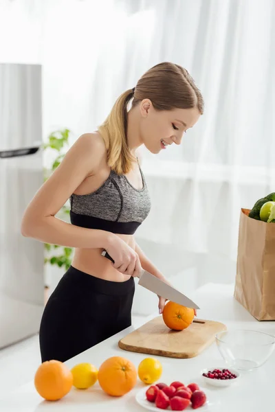 Felice ragazza in abbigliamento sportivo taglio arancione vicino generi alimentari in sacchetto di carta — Foto stock