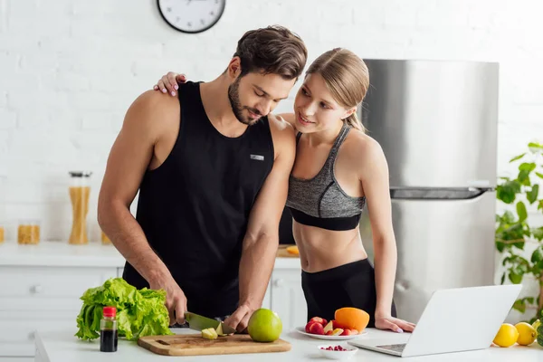 Attractive girl looking at man cutting apple near laptop and fruits in kitchen — Stock Photo