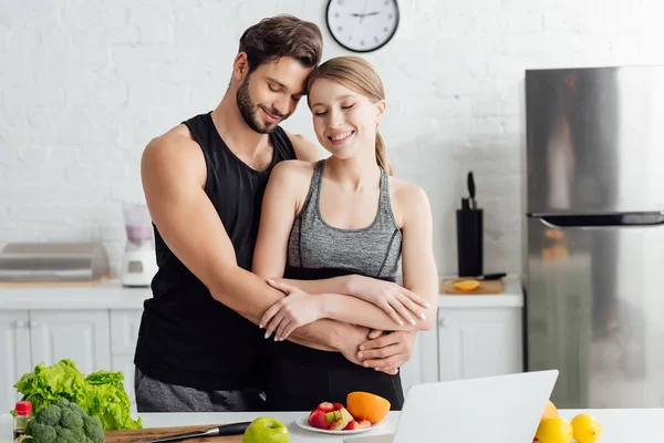 Felice uomo abbracciando ragazza in abbigliamento sportivo vicino laptop e frutta fresca — Foto stock