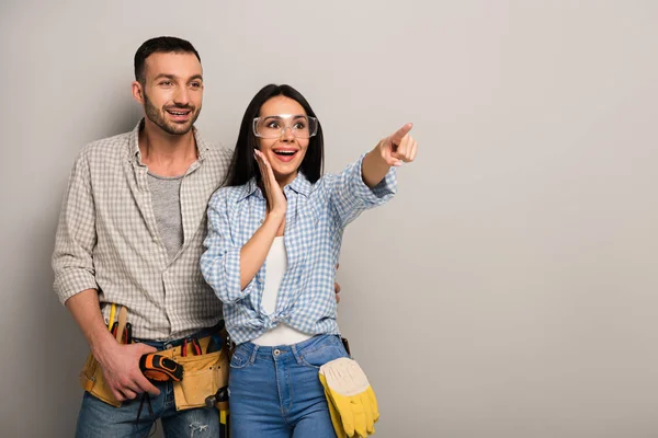 Trabajadores manuales excitados en gafas con cinturón de herramientas apuntando en gris — Stock Photo