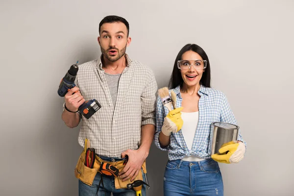 Trabajadores manuales sorprendidos sosteniendo lata de pintura, cepillos y taladro eléctrico en gris — Stock Photo