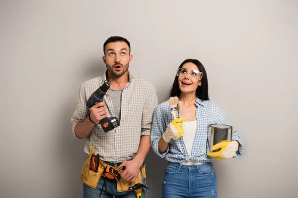 Travailleurs excités tenant la boîte de peinture, brosses et perceuse électrique sur gris — Photo de stock