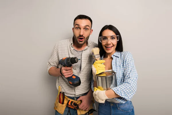 Surprised manual workers holding paint can, brushes and electric drill on grey — Stock Photo