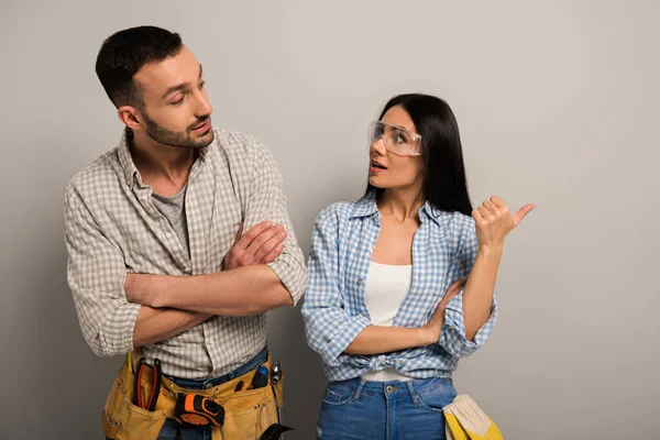 Pensive manual workers in goggles with tool belt pointing on grey — Stock Photo