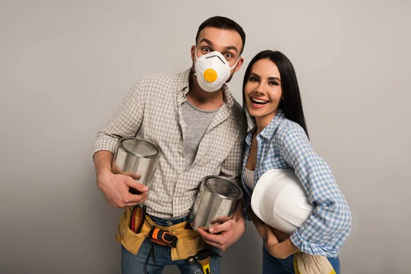 Trabajadores manuales excitados en máscara de seguridad sosteniendo latas de pintura y casco en gris - foto de stock