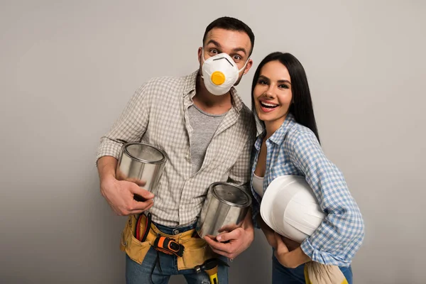 Aufgeregte Arbeiter in Schutzmaske halten Farbdosen und Helm auf grau — Stockfoto