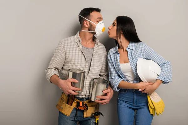 Couple of happy manual workers in safety mask holding paint cans and helmet and kissing on grey — Stock Photo