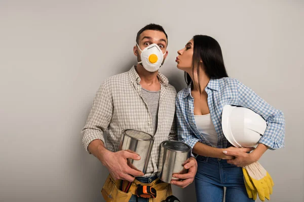 Couple of happy manual workers in safety mask holding paint cans and helmet and kissing on grey — Stock Photo