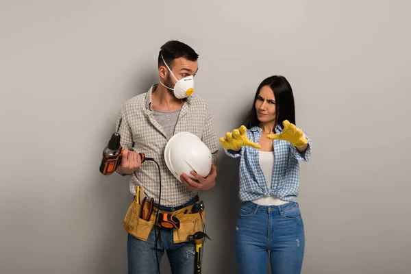 Емоційні ручні працівники в безпечній масці тримають електричний дриль і шолом на сірому — стокове фото