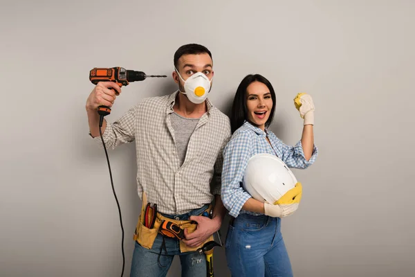 Excited manual workers in safety mask holding electric drill and helmet on grey — Stock Photo