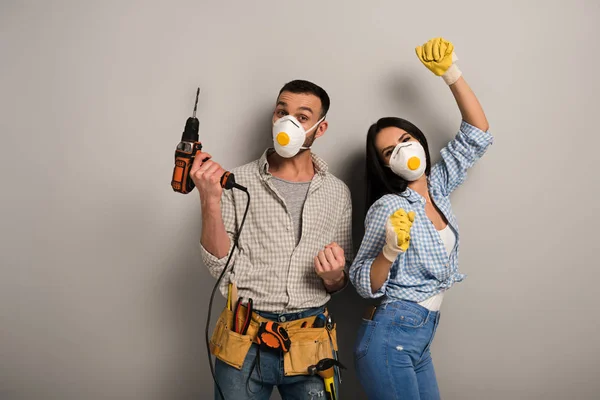 Glückliche Arbeiter in Schutzmasken halten Elektrobohrer auf grau — Stockfoto