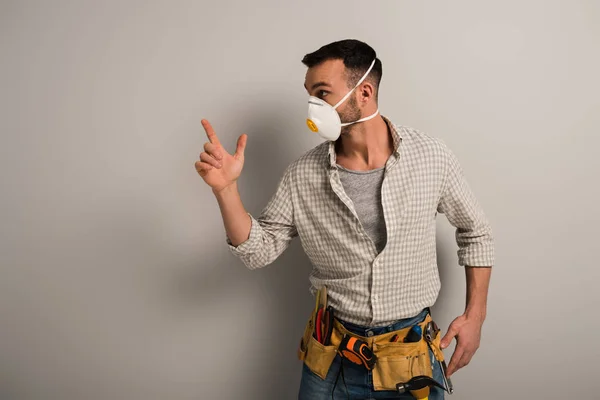 Trabajador manual emocional en máscara de seguridad con cinturón de herramientas haciendo gesto pistola de mano en gris - foto de stock