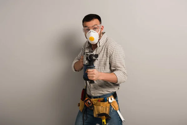 Емоційний майстер в масці безпеки, що тримає електричний дриль на сірому — стокове фото