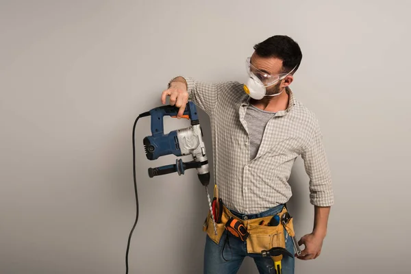 Sorprendido trabajador manual en la máscara de seguridad celebración de taladro eléctrico en gris - foto de stock