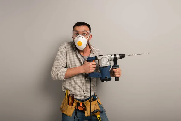 Asustado trabajador manual en la máscara de seguridad celebración de taladro eléctrico en gris - foto de stock